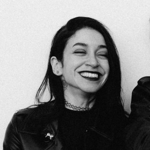 A smiling person with dark hair wearing a black leather jacket and choker, against a plain background.